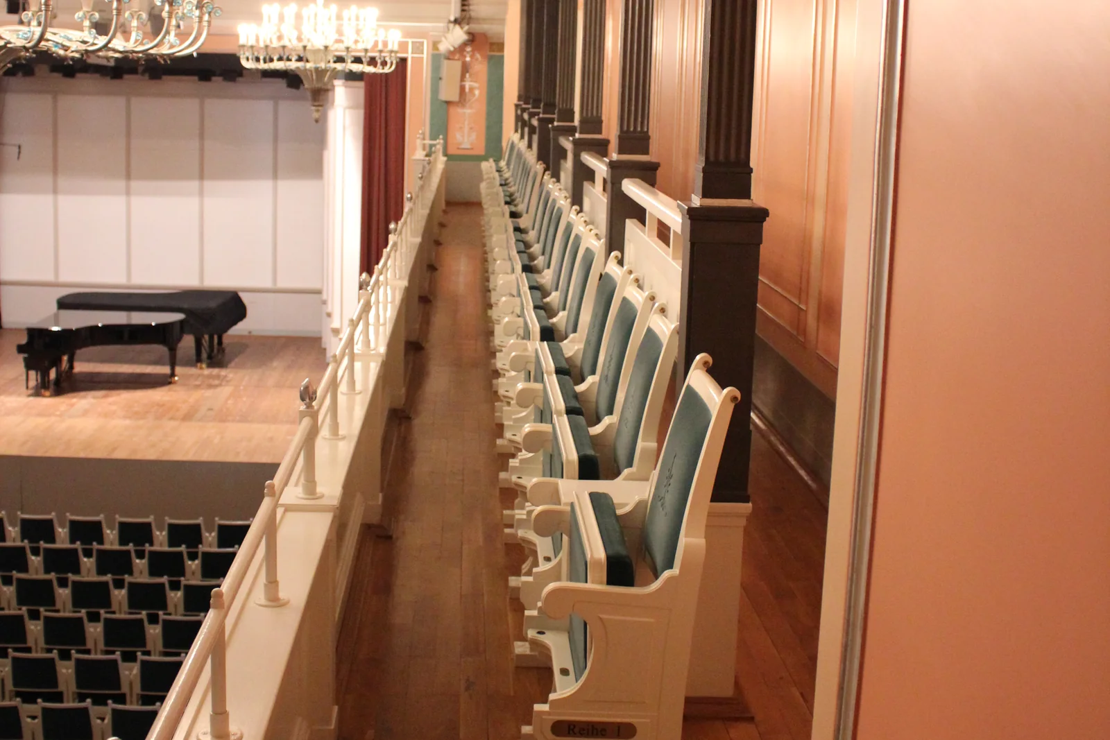 Balconata di una sala da musica: sedute imbottite, pavimento in legno, colonne, palco con pianoforte